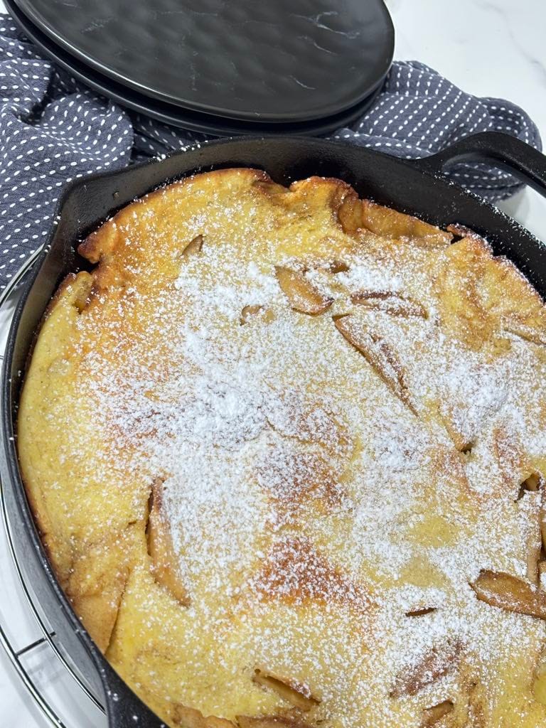 A freshly baked cast-iron apple pancake topped with powdered sugar, golden brown and ready to slice — a rustic German pancake for breakfast.