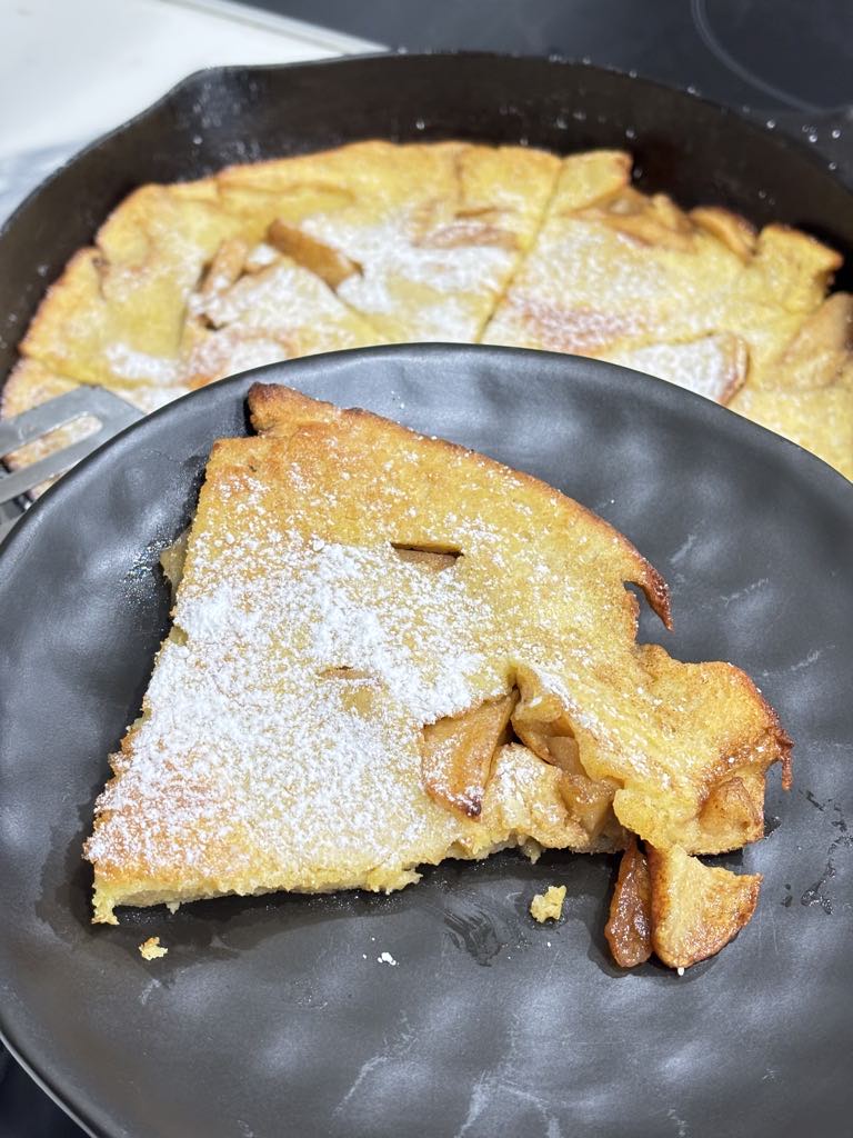 A slice of fluffy cast-iron apple pancake with caramelized apples peeking through, dusted in powdered sugar, served on a black plate.