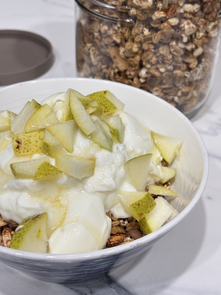 A rustic breakfast bowl featuring Christmas granola with Greek yogurt and diced pear. A large glass jar of homemade granola is visible behind the bowl, showcasing a cozy, festive morning vibe.