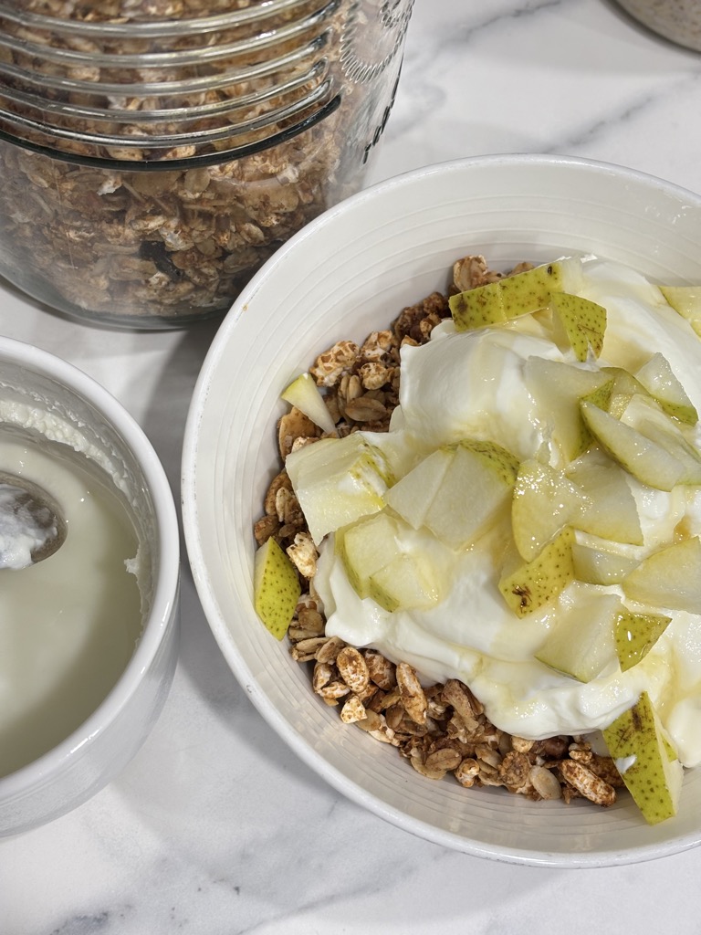 A top-down shot of a homemade granola bowl, featuring yogurt and fresh pear, with a large airtight jar of gingerbread granola beside it. A cozy, wholesome breakfast idea.