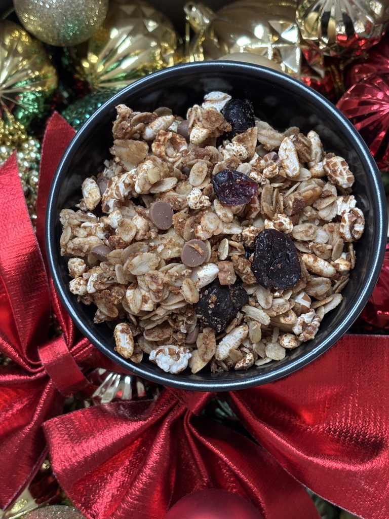 A black bowl filled with Christmas gingerbread granola, dotted with chocolate chips and dried cranberries, surrounded by red bows and shiny ornaments. A perfect homemade gift idea for the holidays.