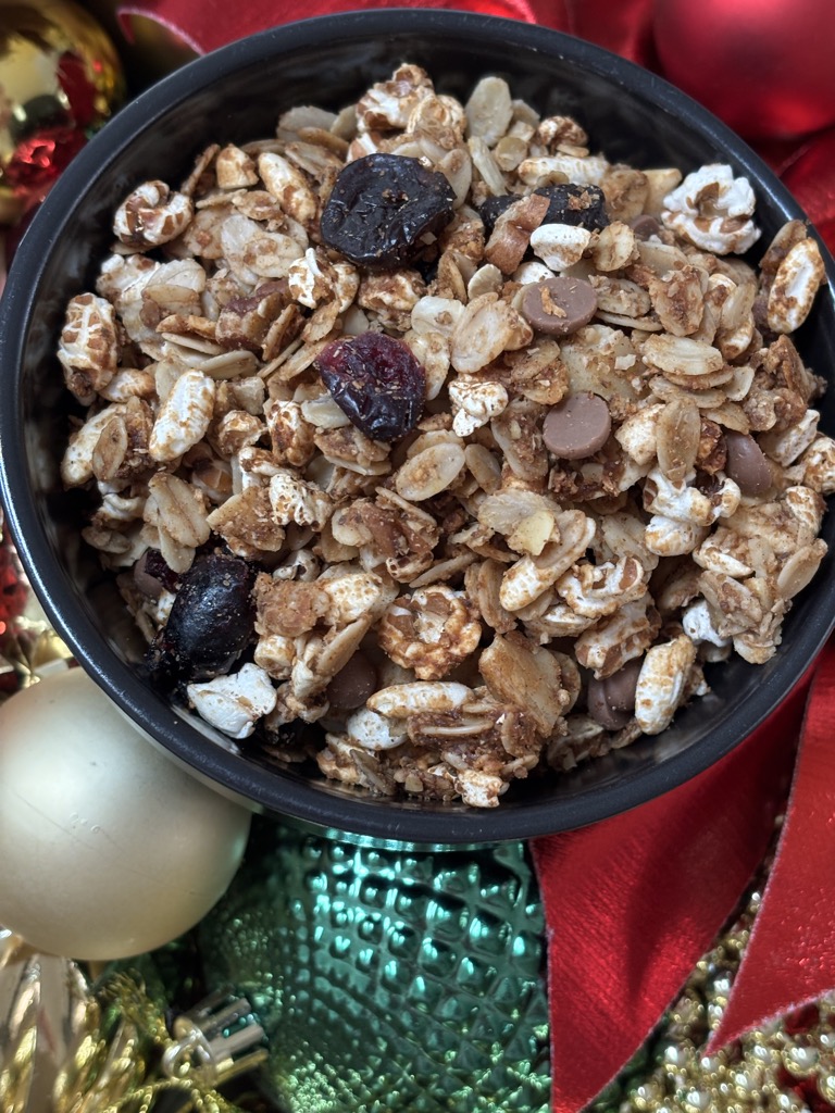 A black bowl filled with homemade gingerbread granola, chocolate chips, and dried cranberries, surrounded by red and gold Christmas ornaments. Perfect for holiday gifting or easy seasonal breakfasts.