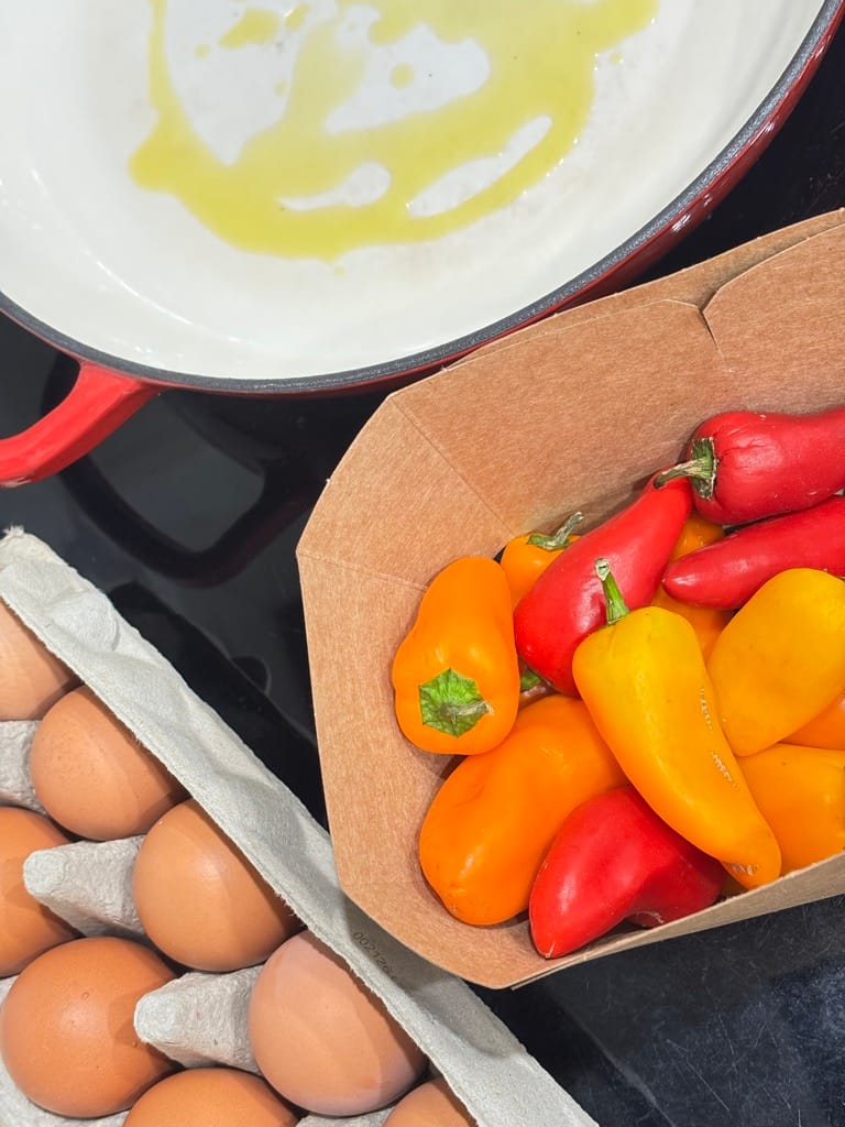 A rustic kitchen setup with a carton of brown eggs, a basket of colorful red, yellow, and orange bell peppers, and olive oil drizzled in a red skillet — the fresh start of a traditional shakshuka recipe.