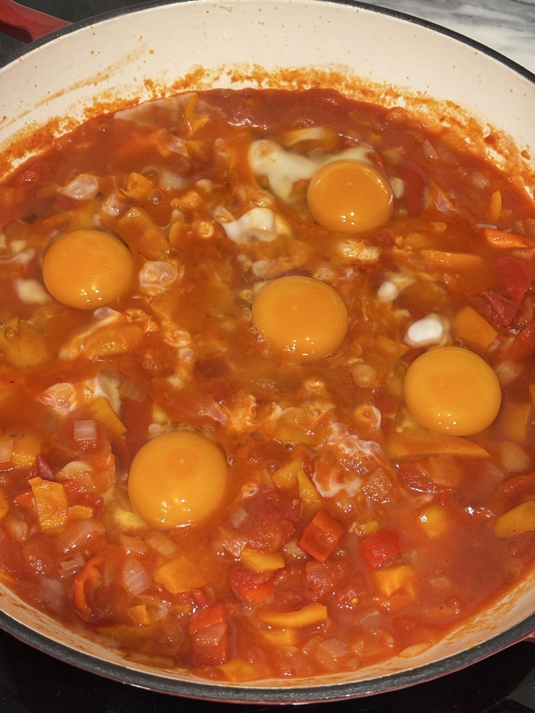 A pan of simmering tomato and pepper sauce with fresh eggs just added, their bright yellow yolks contrasting the deep red sauce — a comforting North African tomato dish in progress.
