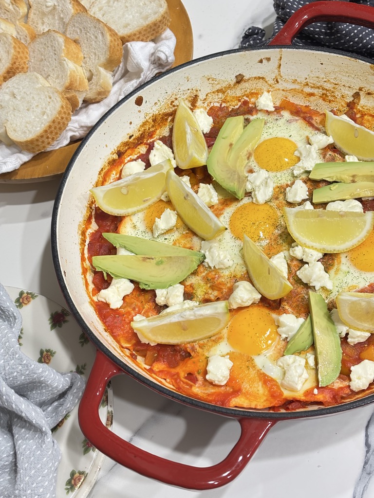 A beautiful finished shakshuka in a red skillet, topped with feta cheese, creamy avocado slices, and lemon wedges — ready to serve with crusty bread.