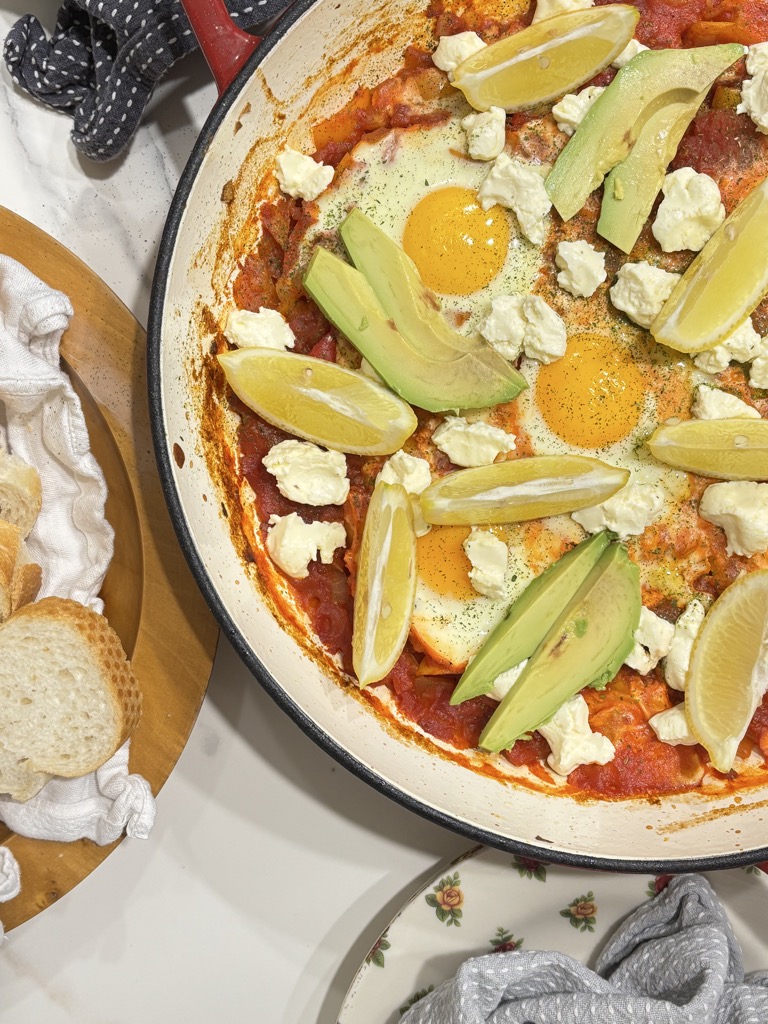 A hearty North African shakshuka in a red enamel skillet, garnished with avocado slices and fresh herbs — a comforting and colorful tomato-based breakfast.