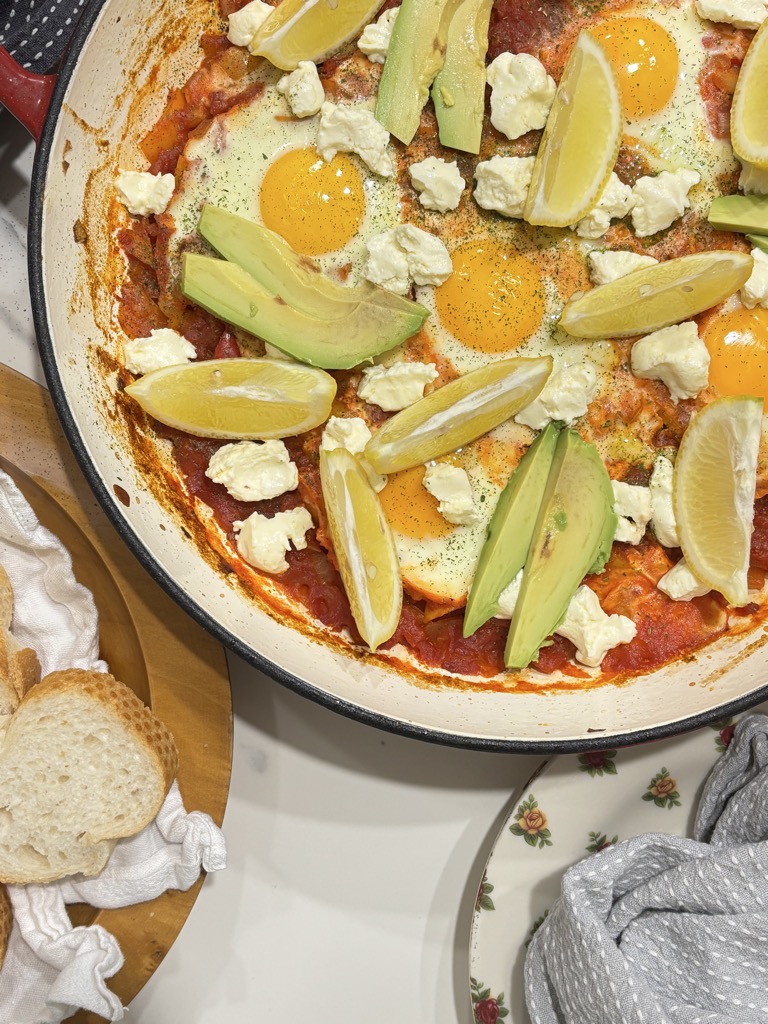 A hearty North African shakshuka in a red enamel skillet, garnished with avocado slices and fresh herbs — a comforting and colorful tomato-based breakfast.
