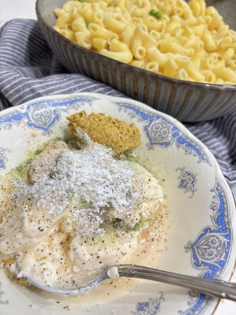 A close-up bowl of mayonnaise, mustard, spices, and vinegar being mixed to create a creamy dressing for a traditional macaroni salad, with a dish of cooked elbow macaroni in the background.