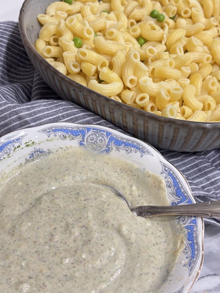 A bowl of fully mixed, speckled creamy dressing sitting beside a dish of cooked elbow macaroni with peas, ready to be combined for classic macaroni salad.