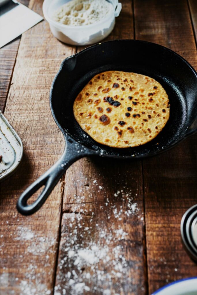 A golden, blistered flatbread cooks in a cast iron skillet on a flour-dusted wooden countertop. The rustic setup, complete with a bowl of flour and baking tools, highlights traditional home cooking and the durability of cast iron pans for everyday recipes.
