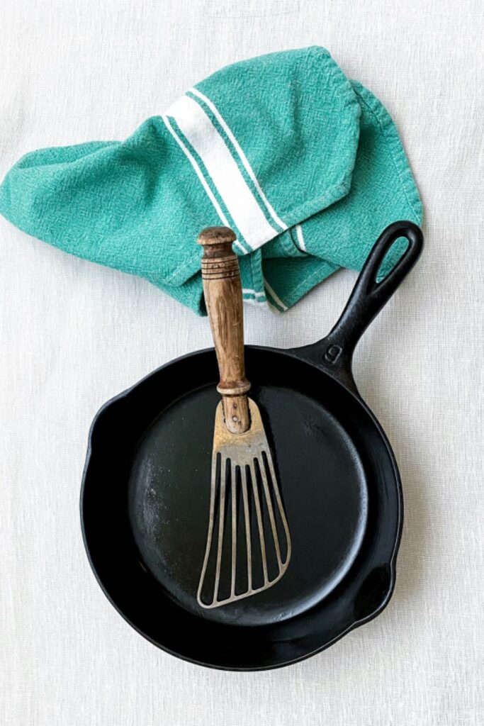 A clean cast iron skillet rests on a white cloth alongside a vintage wooden-handled spatula and a folded green tea towel. The image showcases simple, old-school kitchen tools and a well-maintained cast iron pan ready for cooking.