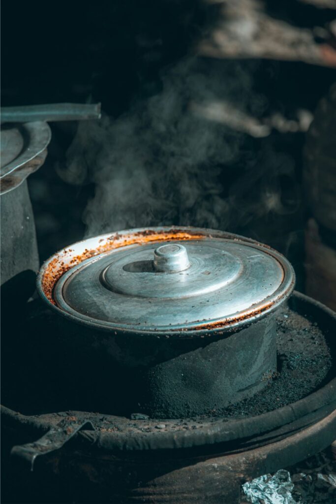 A weathered cast iron dutch oven with a metal lid simmers over an outdoor fire pit, with steam rising through the edges. The pot shows signs of long-term use, capturing the rugged charm and resilience of cast iron cookware in traditional cooking settings.
