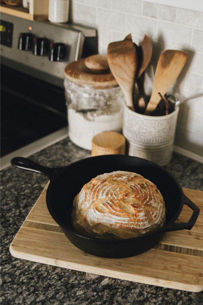 A round artisan loaf with a beautifully floured spiral crust sits in a black cast iron dutch oven on a wooden board. The warm kitchen setting, complete with wooden utensils and jars in the background, highlights the reliability of cast iron for baking crusty homemade bread.