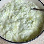 A bowl of creamy cucumber salad with sliced cucumbers coated in a thick white dressing, served in a simple bowl with a spoon.