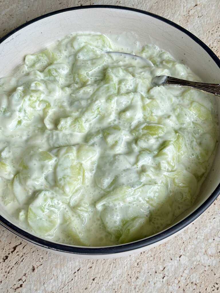 A bowl of creamy cucumber salad with sliced cucumbers coated in a thick white dressing, served in a simple bowl with a spoon.