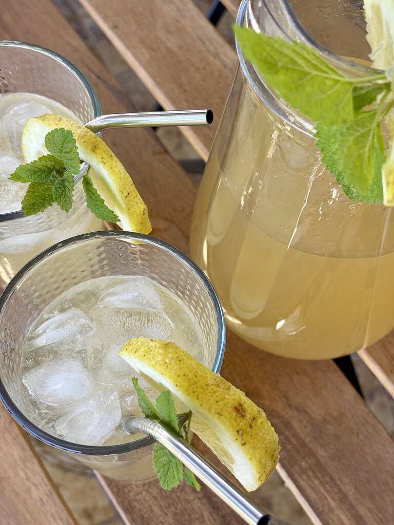 Old-fashioned lemonade recipe served over ice in two glasses with fresh mint and lemon slices, next to a glass pitcher on a wooden outdoor table.