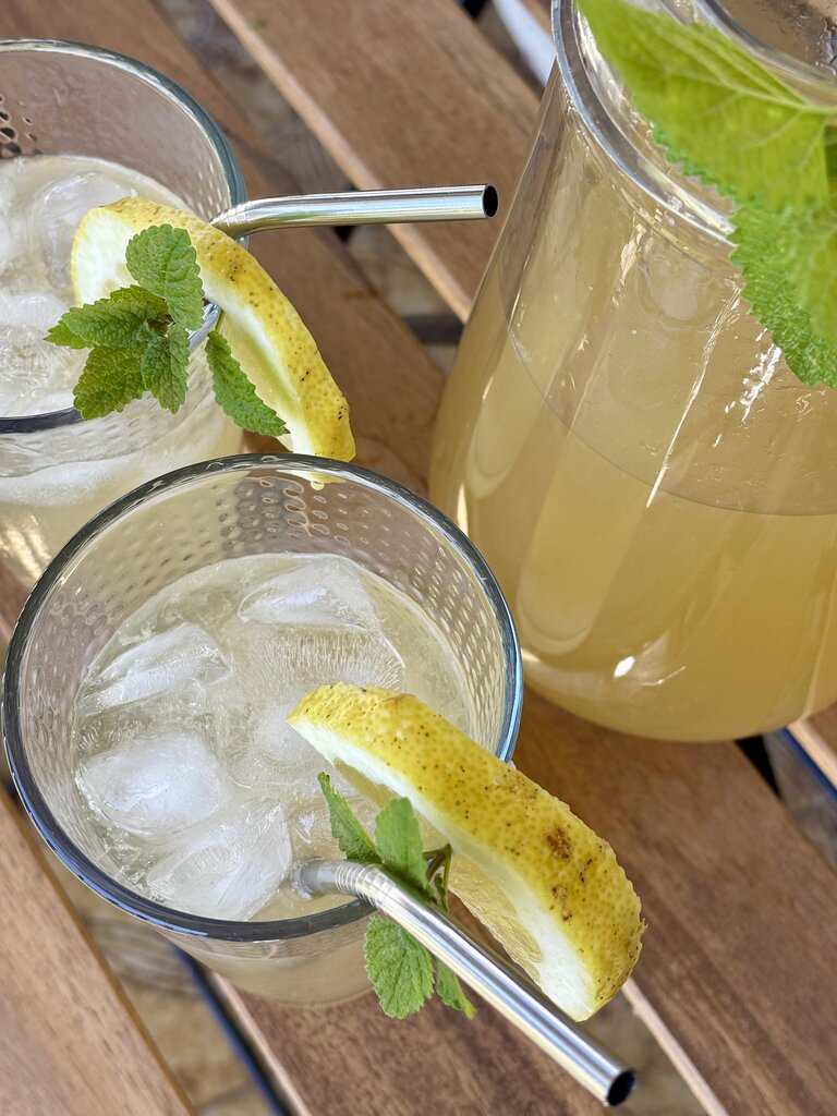 Homemade old-fashioned lemonade recipe poured over ice in textured glasses with lemon wedges and mint sprigs on a rustic wooden table.