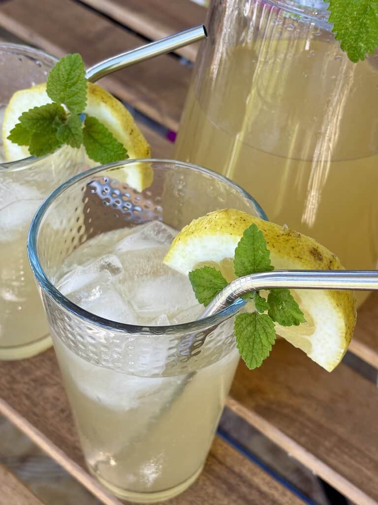 Close-up of a classic old-fashioned lemonade recipe from scratch served in a glass with ice cubes, fresh lemon wedge, mint leaves, and a reusable metal straw.