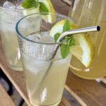 Easy old-fashioned lemonade recipe served chilled over ice with fresh lemon slices and mint, styled outdoors on a wooden table with a glass pitcher in the background.