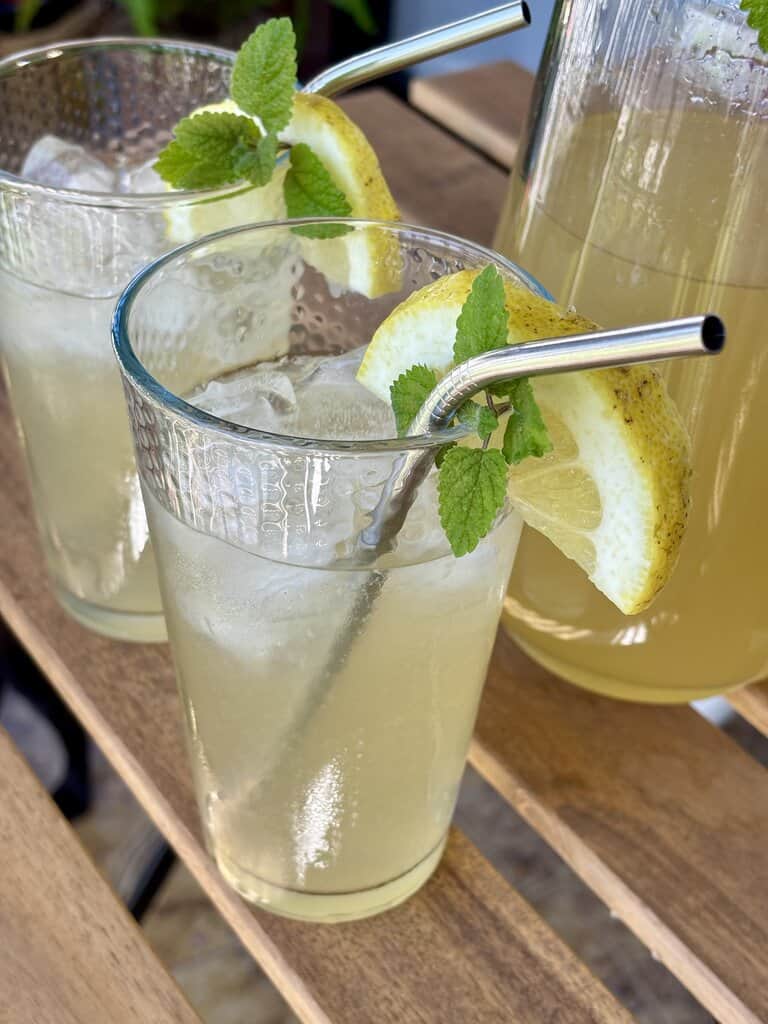 Easy old-fashioned lemonade recipe served chilled over ice with fresh lemon slices and mint, styled outdoors on a wooden table with a glass pitcher in the background.