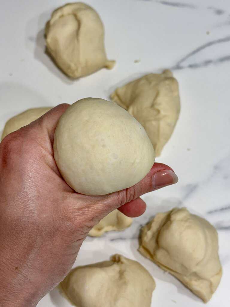 Hand holding a smooth dough ball while shaping homemade soft dinner rolls on a work surface.